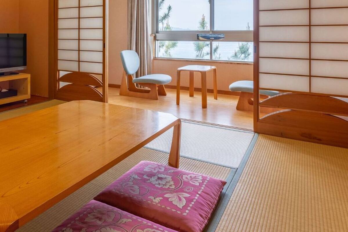 A traditional Japanese room with tatami flooring, a low wooden table, a television on a stand, and a sliding door with a view of the outdoors.