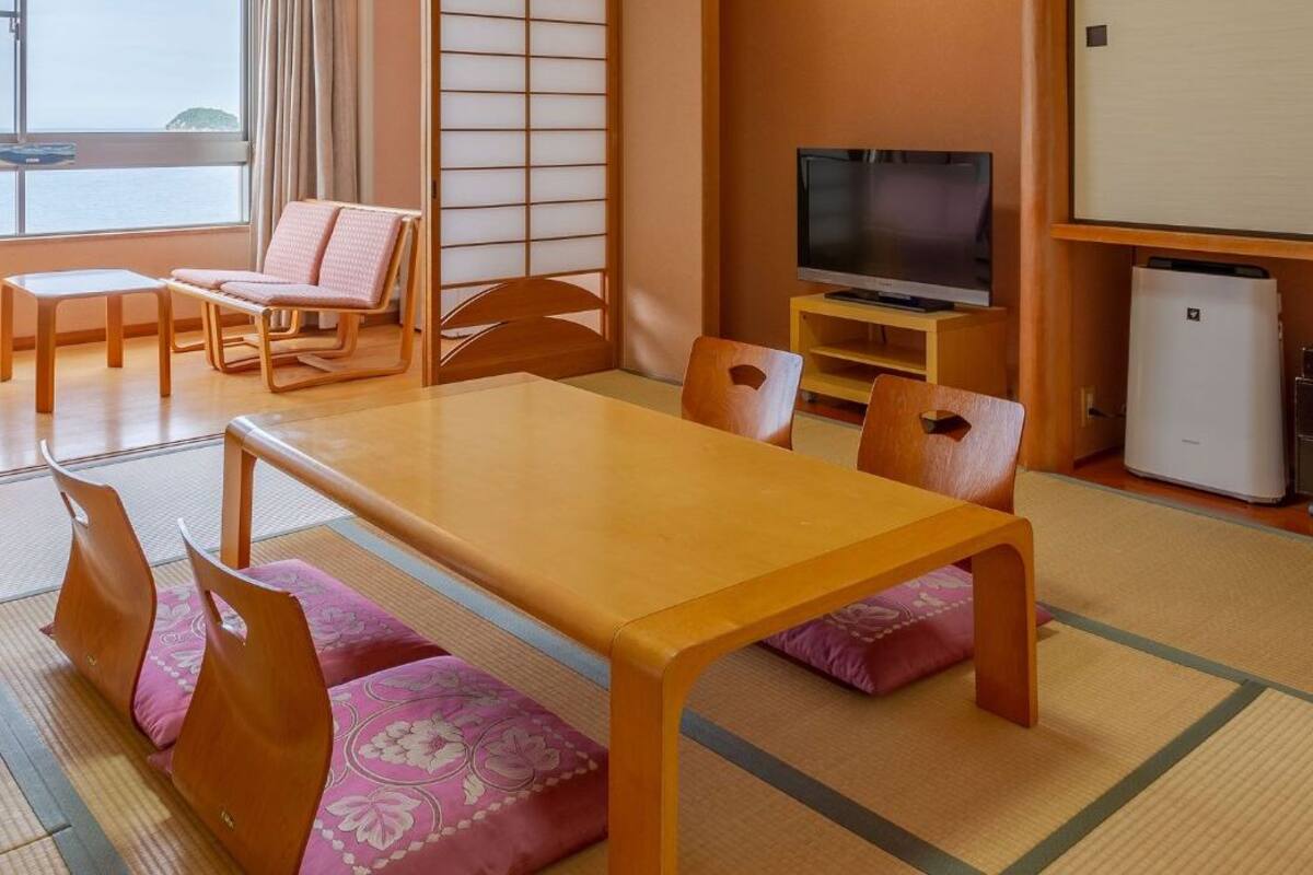 A traditional Japanese-style room with tatami flooring, a low table, wooden chairs, a television, and a refrigerator.