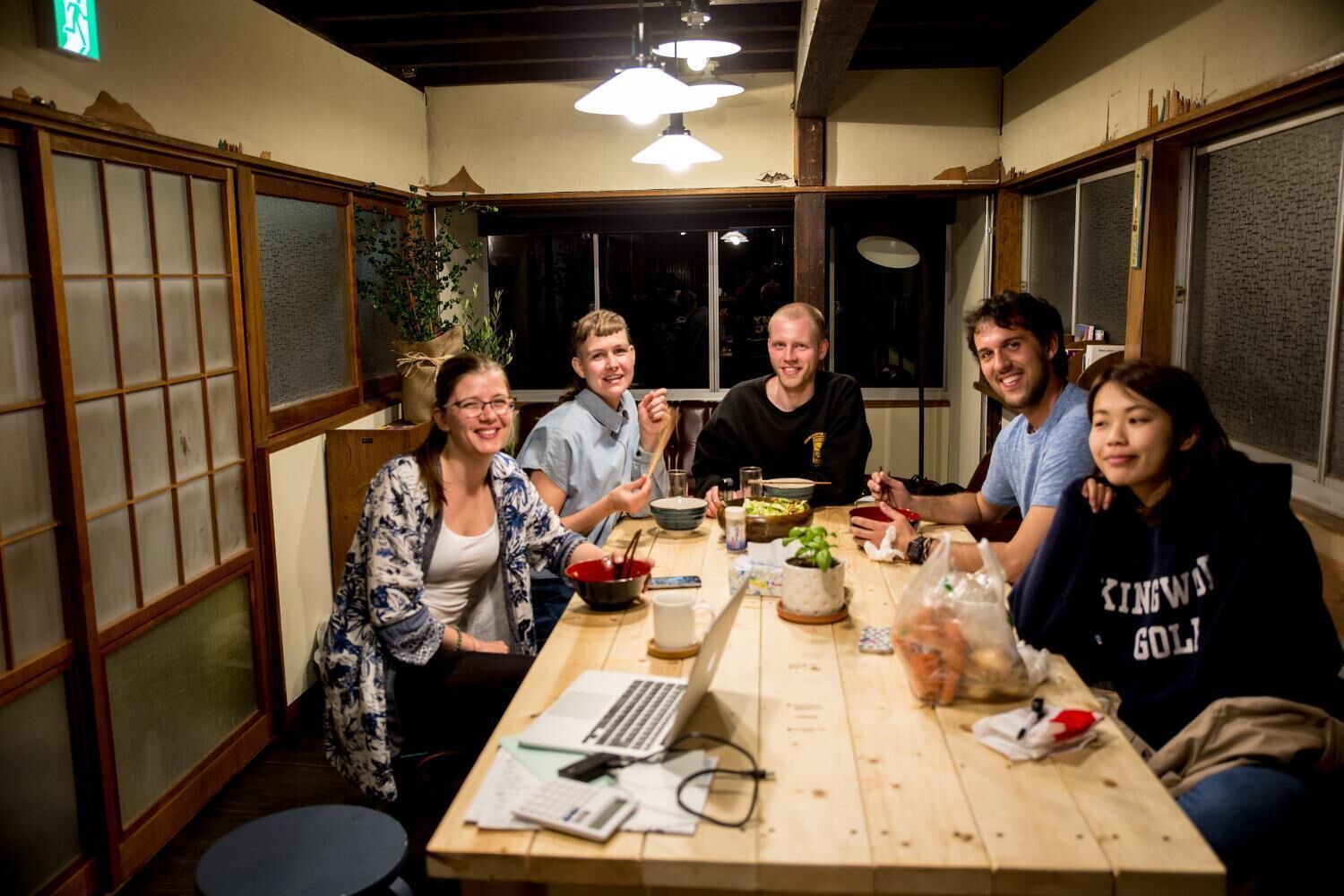 A group of people sitting around a wooden table with food and drinks, in a room with traditional Japanese sliding doors and a ceiling fan.