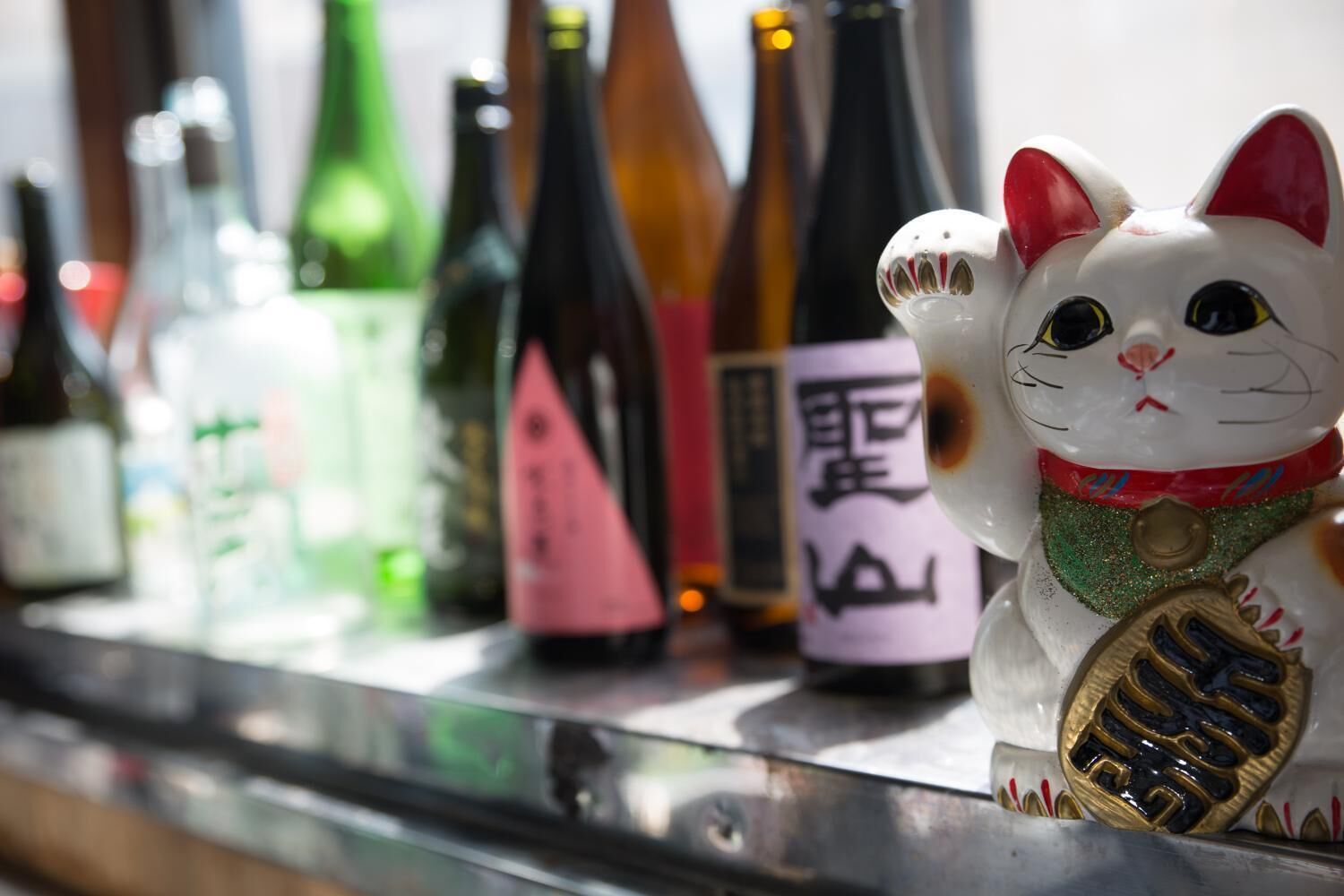Eine Auswahl an Flaschen mit Etiketten, darunter eine mit japanischen Schriftzeichen, sowie eine dekorative Katzenfigur mit Medaille.