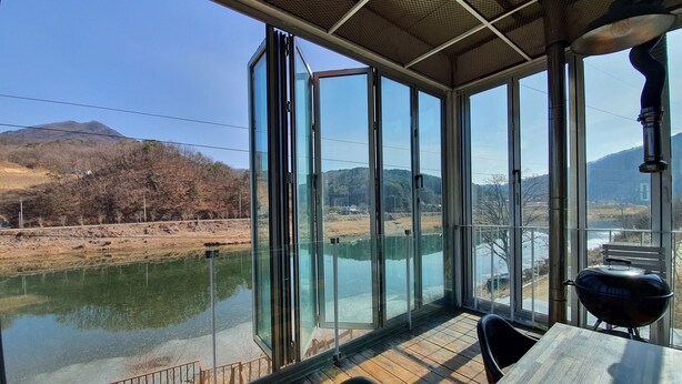Habitación con amplias ventanas que dan a un río y colinas. Cuenta con una terraza de madera en el exterior.