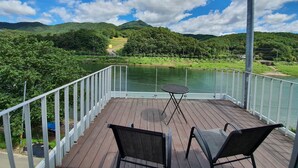 Una terraza de madera con dos sillas y una mesita, con vistas a un río y colinas verdes.