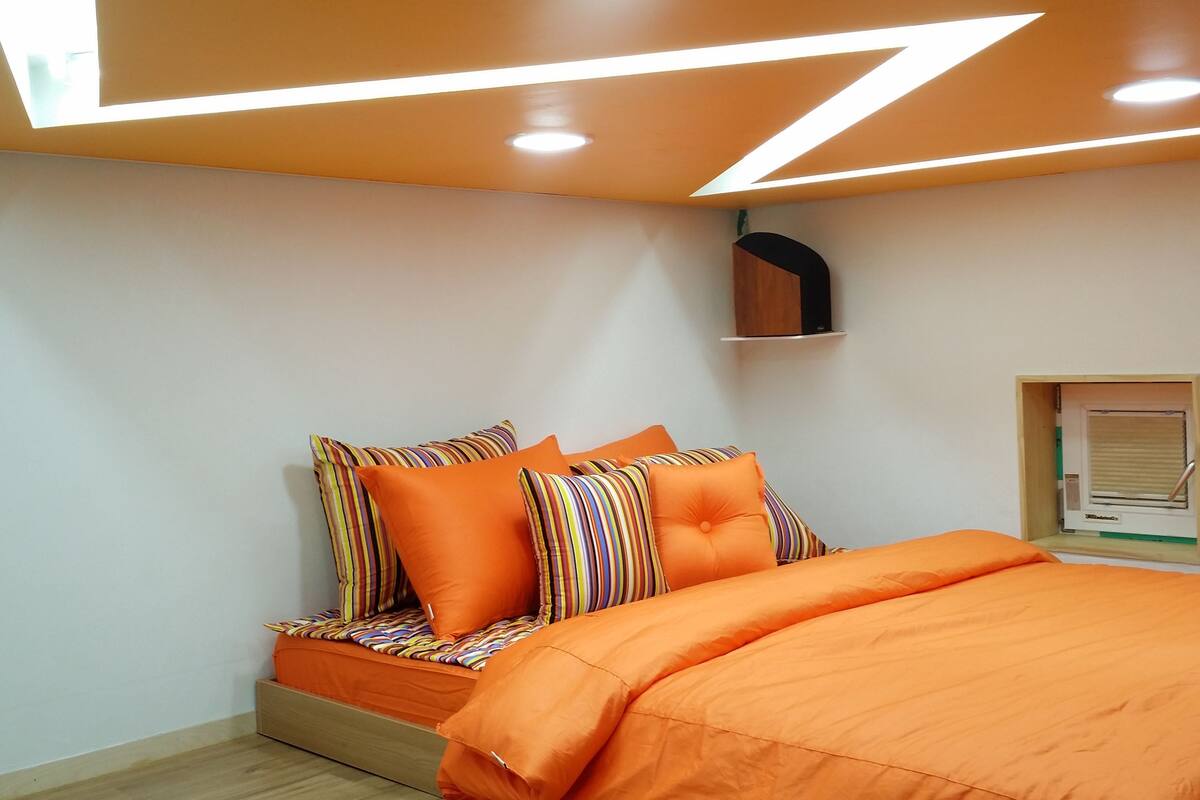 Ein modernes Schlafzimmer mit Bett, orangefarbener Bettwäsche und einem besonderen Design der Decke.