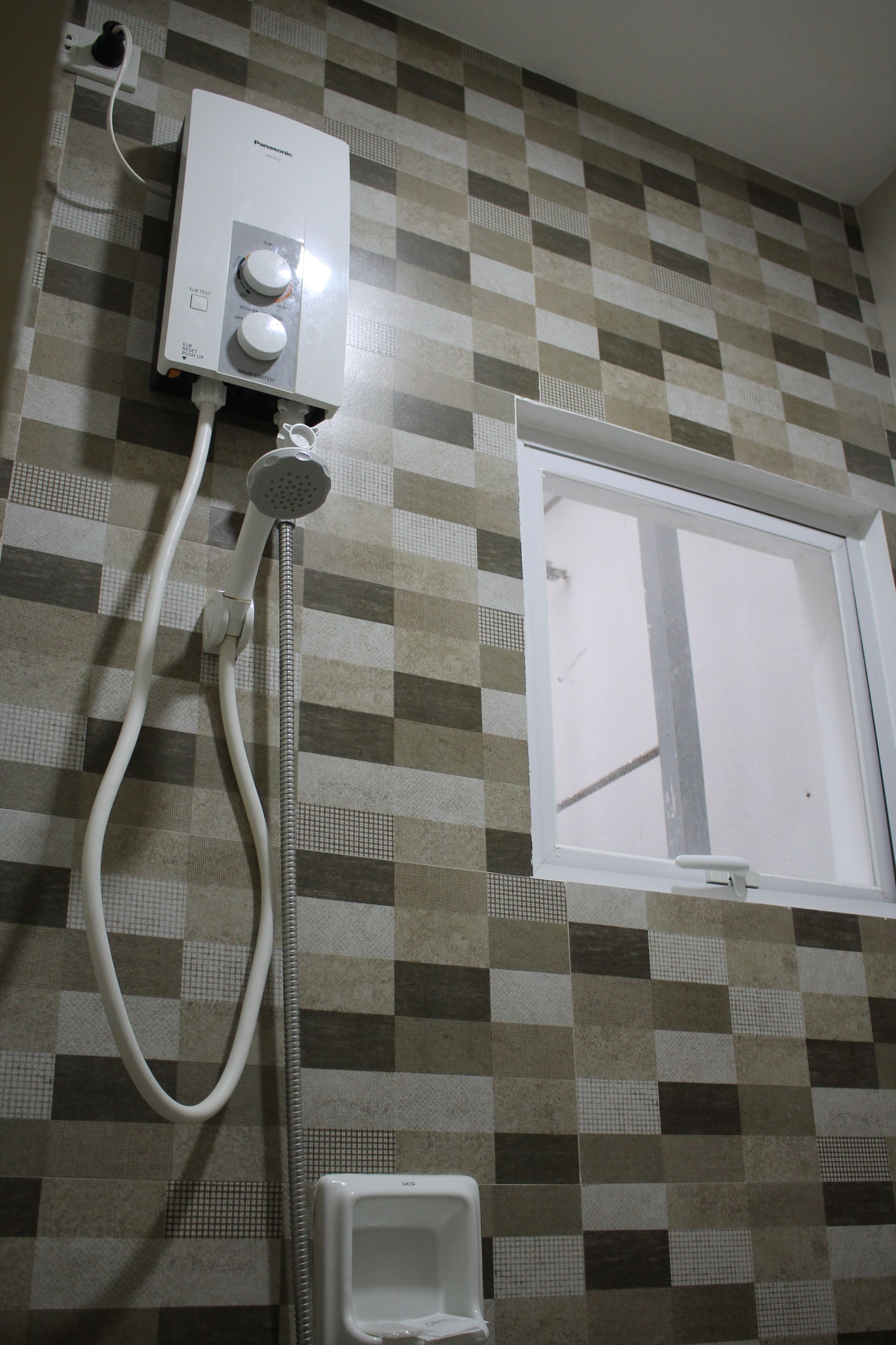 Un baño con pared de baldosas a cuadros, un calentador de pared, un cabezal de ducha manual y una ventana.