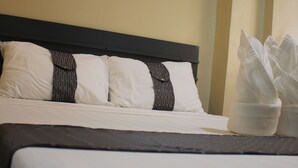Una cama bien hecha con almohadas blancas y oscuras, un cuadro de un paisaje urbano en la pared, y una cesta blanca para toallas al lado.