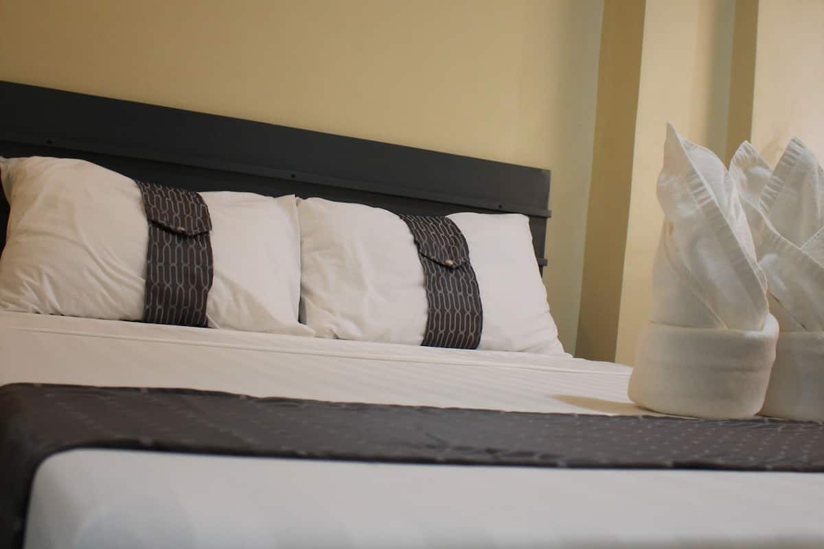 A neatly made bed with white and dark pillows, a painting of a cityscape on the wall, and a white towel basket on the side.