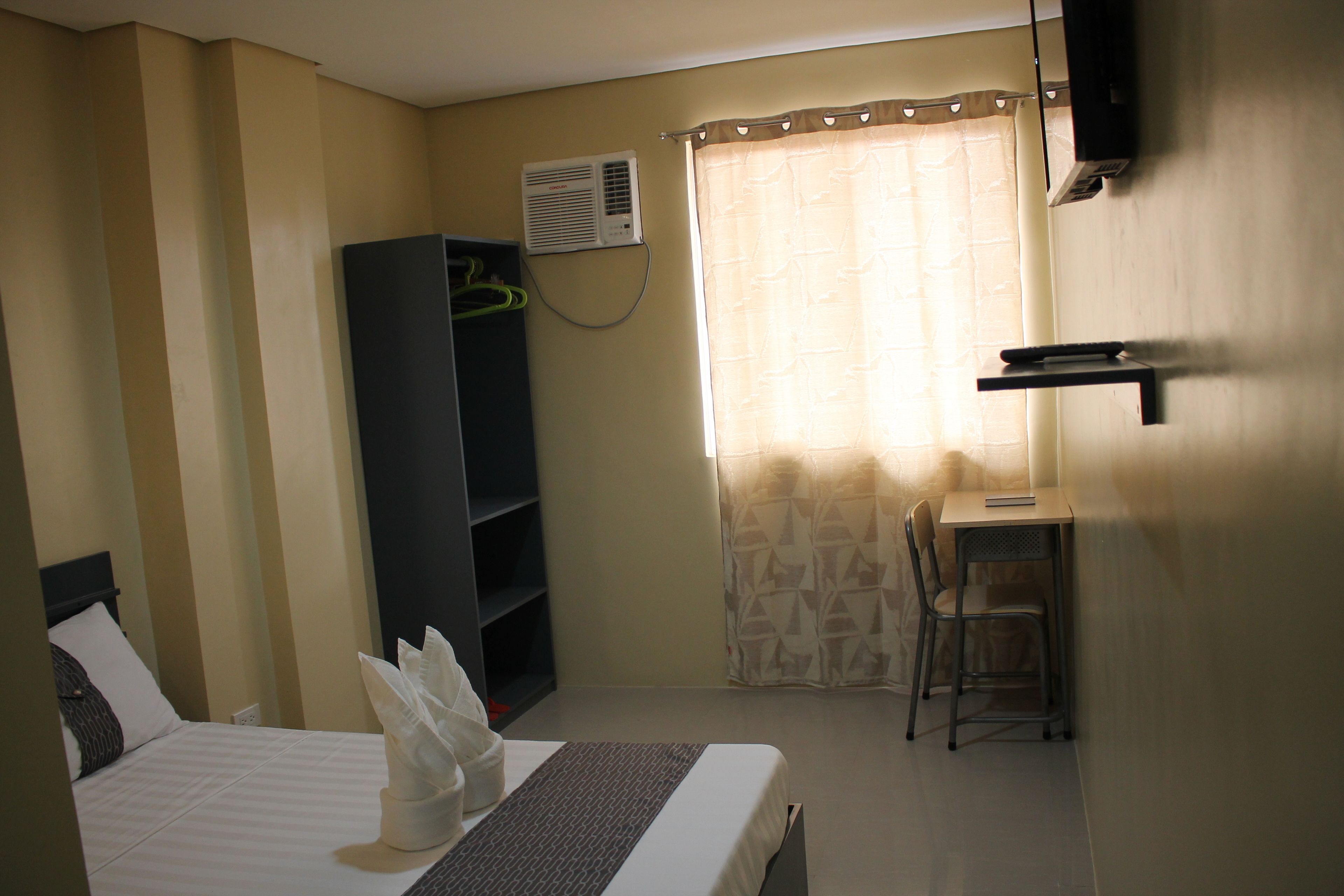 Una habitación de hotel con cama, escritorio, silla, armario y ventana con cortinas.