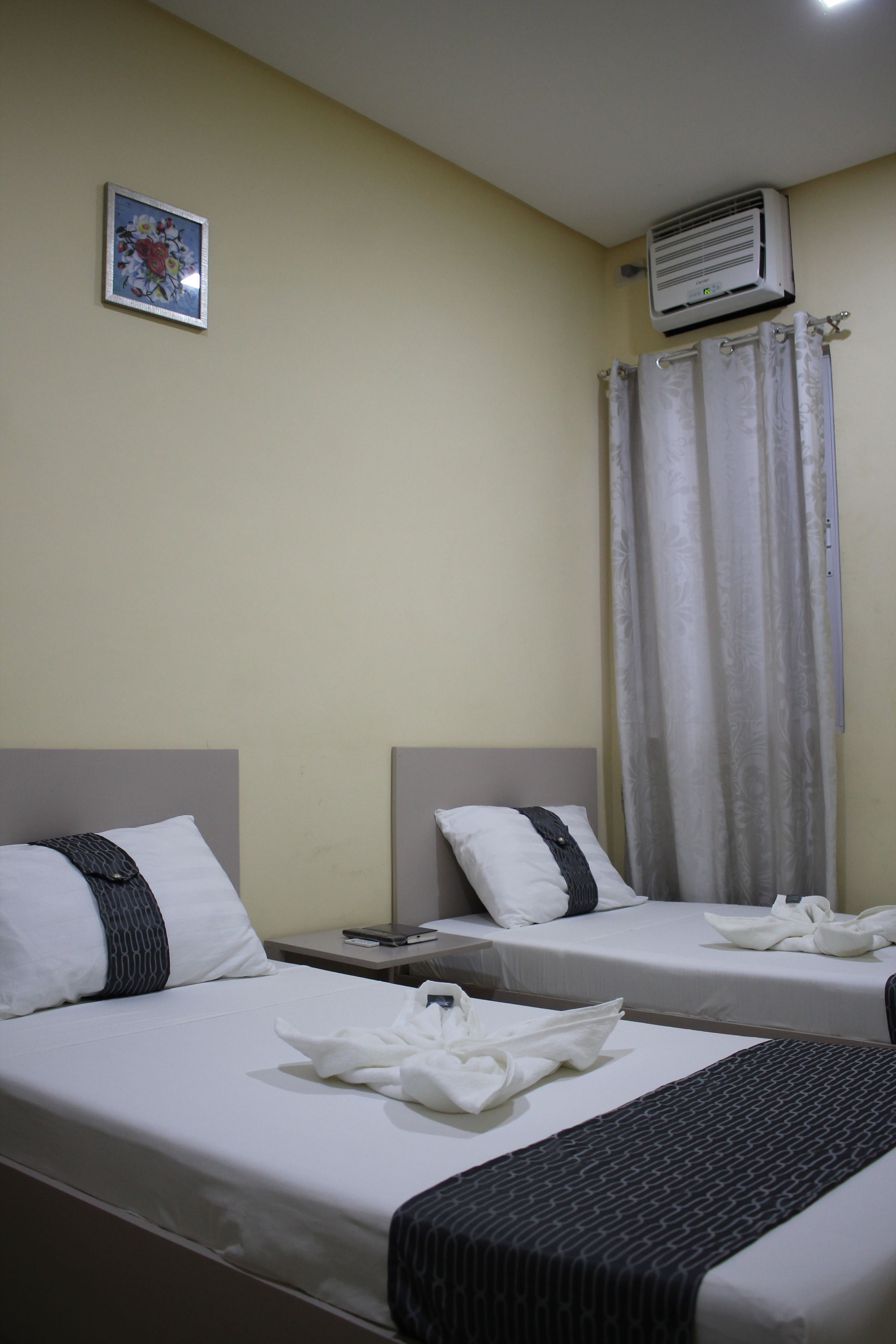 Two single beds with white linens and dark headboards, a small bedside table with a lamp, and a wall-mounted air conditioner.