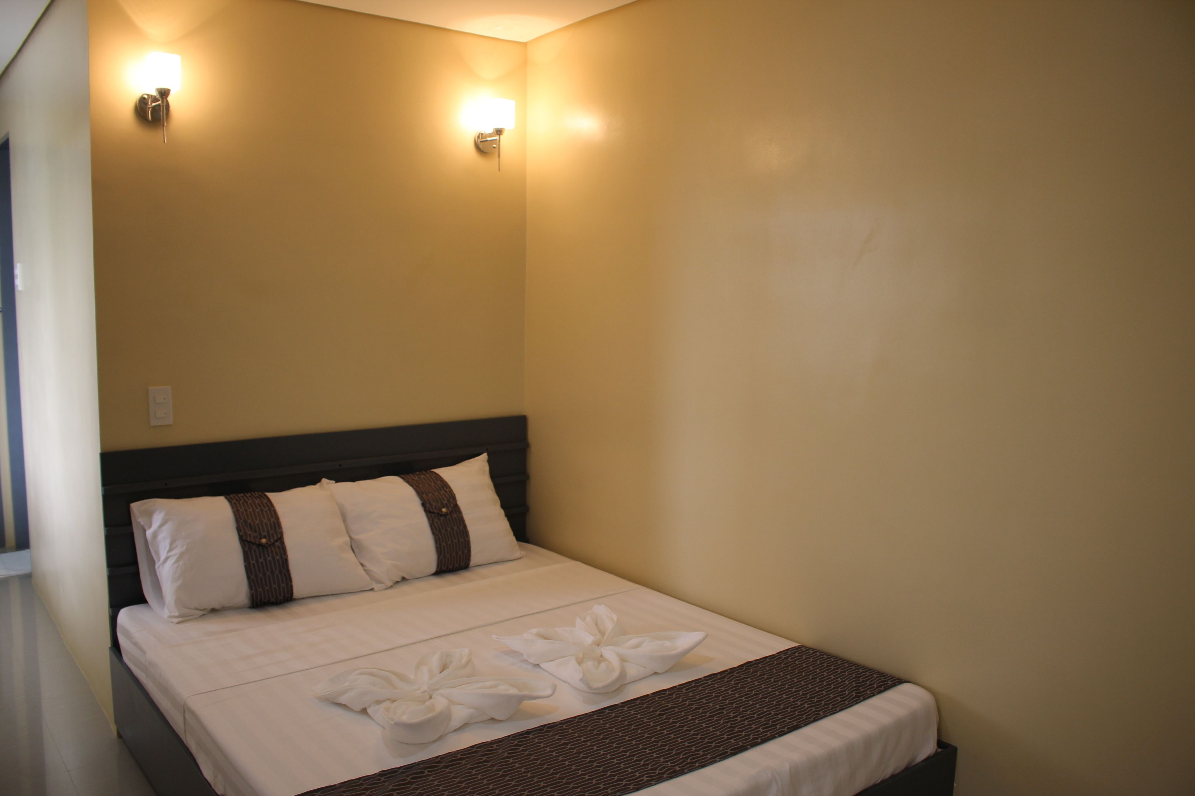 Una cama bien hecha con ropa de cama blanca y detalles marrones, dos lámparas fijadas a la pared y una pared lisa.