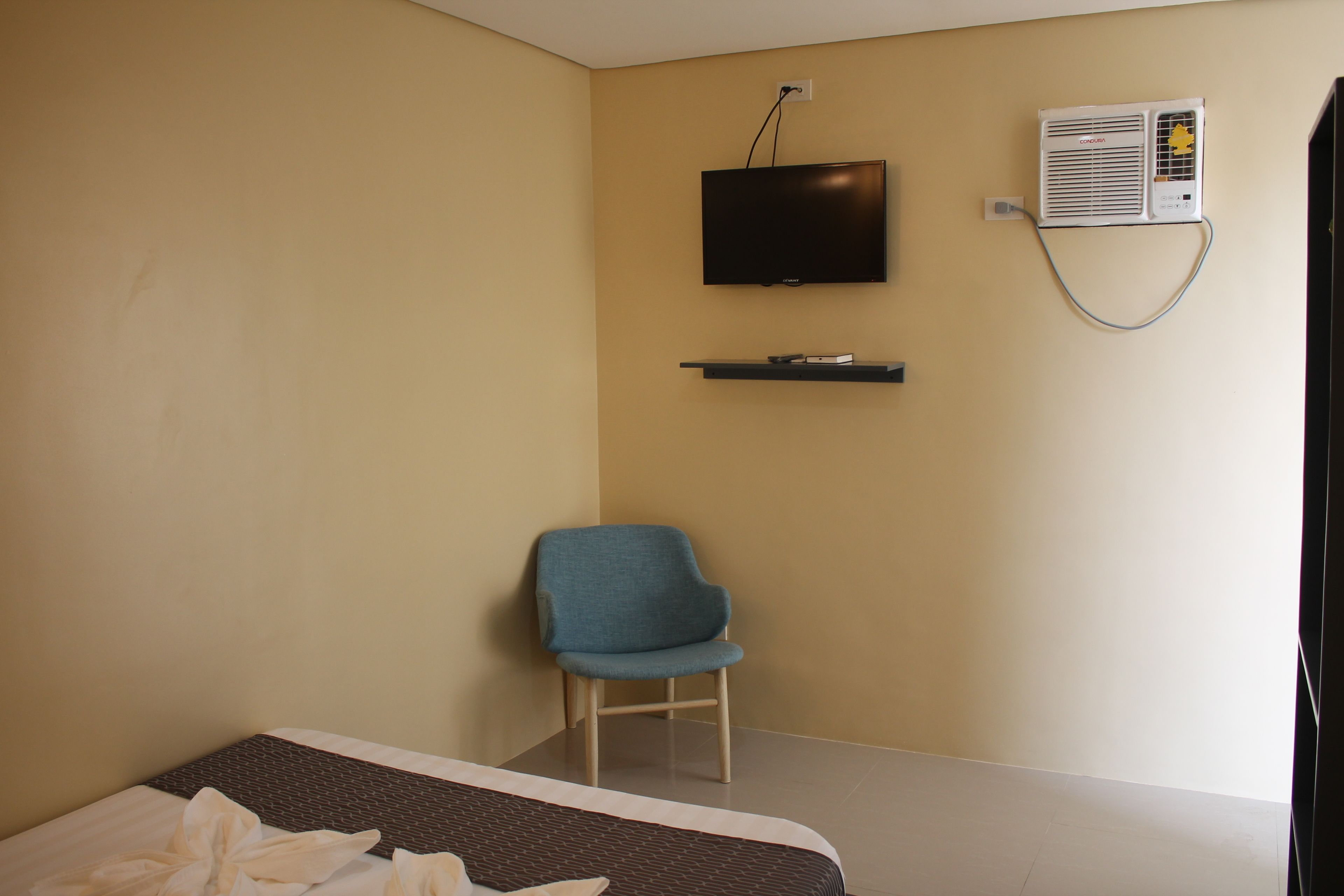 A hotel room with a bed, a blue chair, a flat-screen TV, and an air conditioning unit.