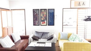 A living room with a brown leather sofa, a yellow armchair, and a black coffee table with a magazine and a pen. There are three framed artworks on the wall and a wooden cabinet with hanging items. - Condominio Mozu 102 (Sakai)