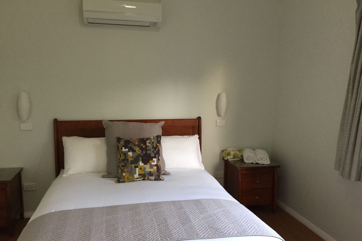 A hotel room with a large bed, a wooden headboard, a bedside table with towels, and an air conditioning unit on the wall.
