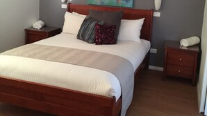 Een netjes opgemaakt bed met een houten hoofdeinde, twee nachtkastjes en een kleurrijk abstract schilderij aan de muur.