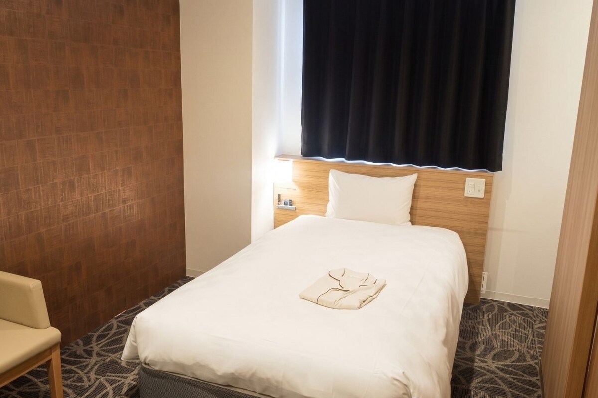Una habitación de hotel con una cama, una silla, un ventanal con cortinas y una alfombra estampada.