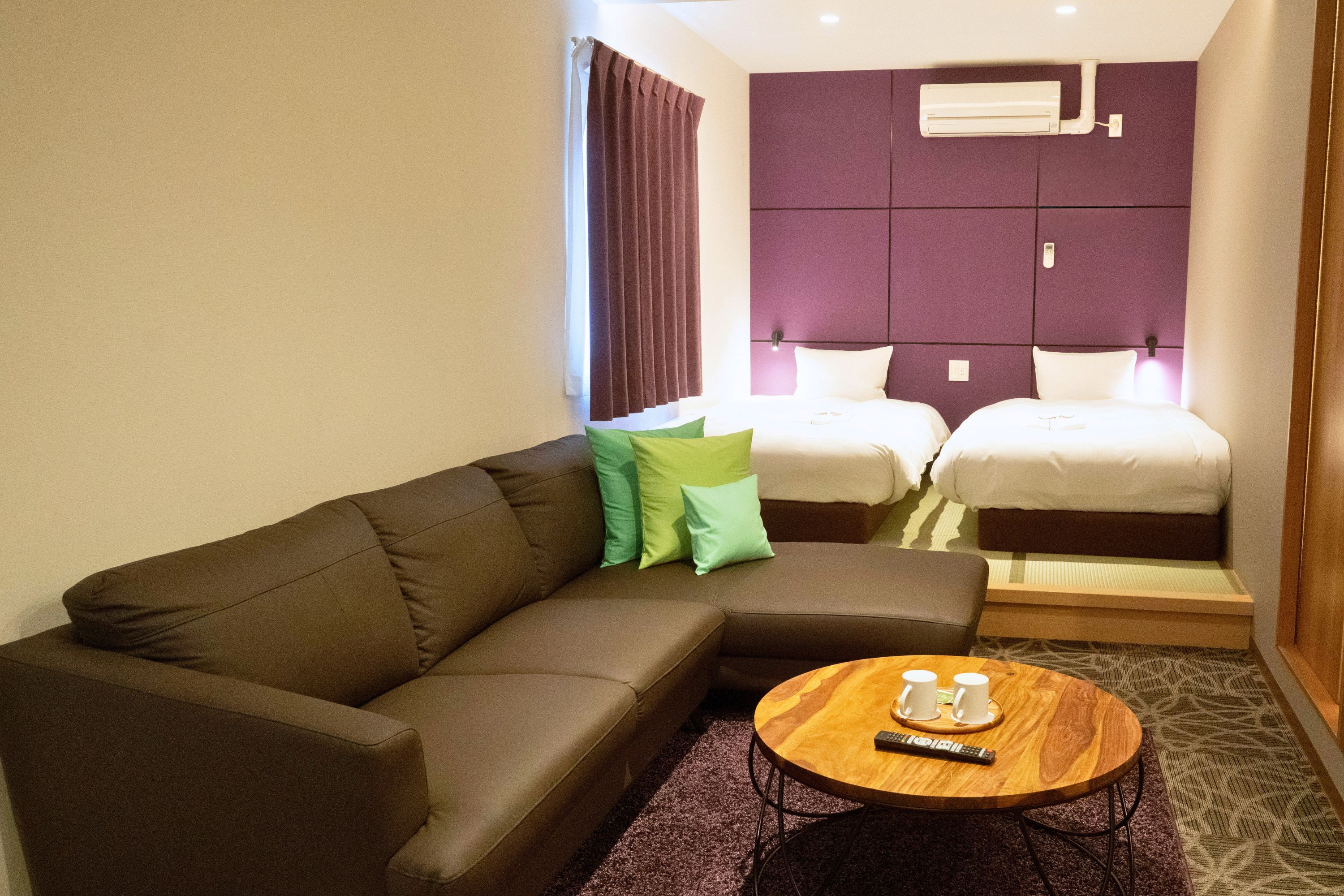 A hotel room with a sofa, two beds, a round wooden coffee table, and an air conditioning unit on the wall.