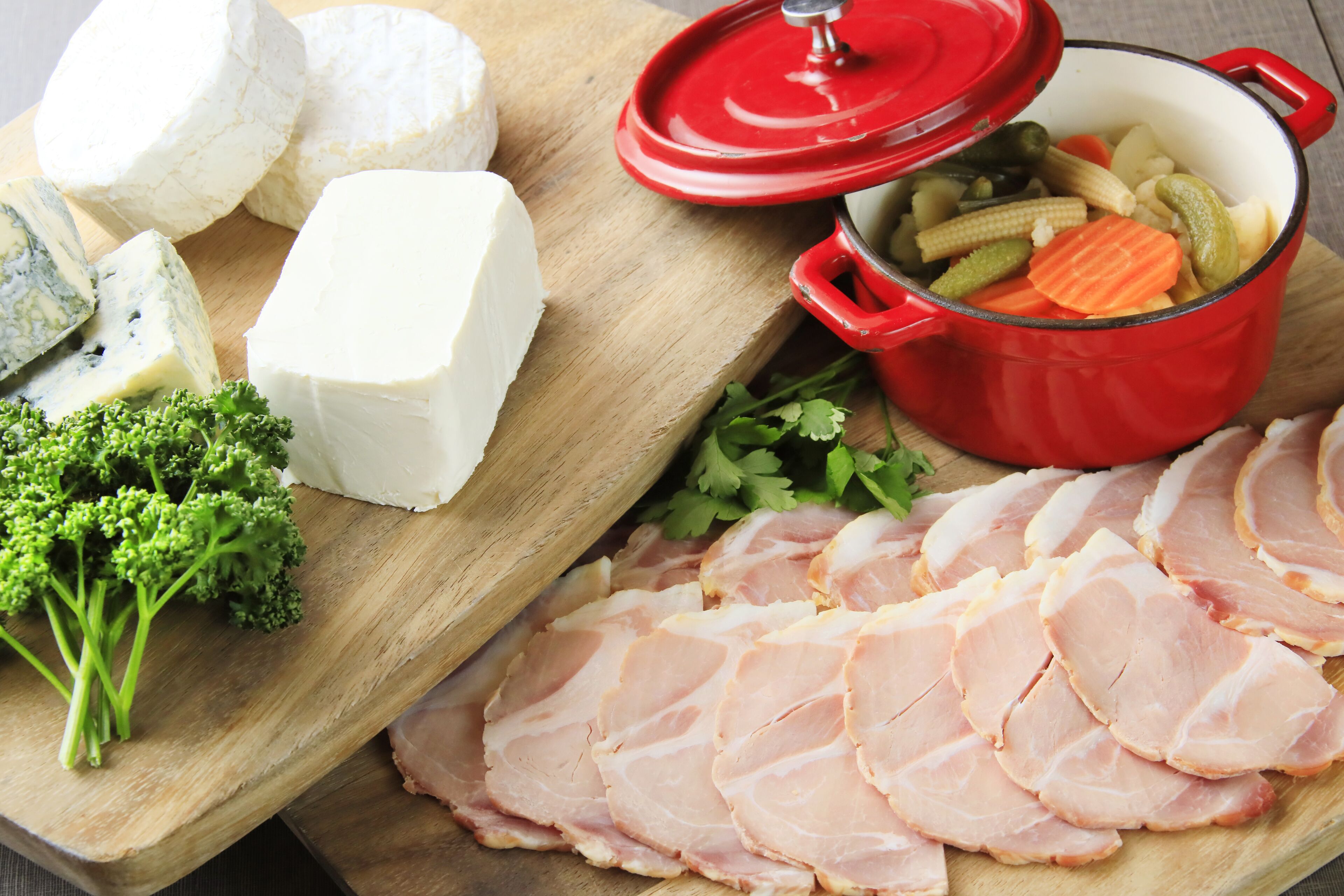 A wooden cutting board with various cheeses, sliced ham, and a red pot with vegetables.