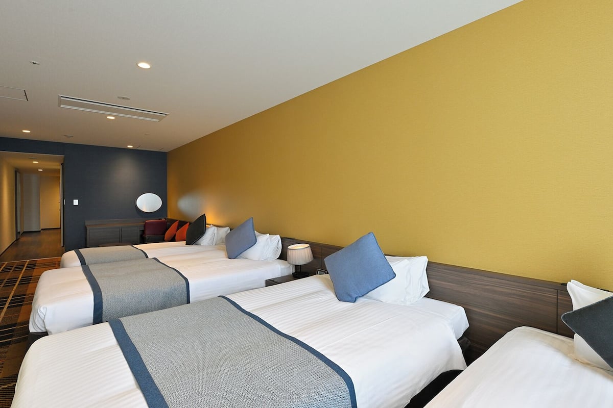 Ett hotellrum med fyra sängar placerade i en rad, en hall som leder till ett annat rum och en väggmonterad TV.