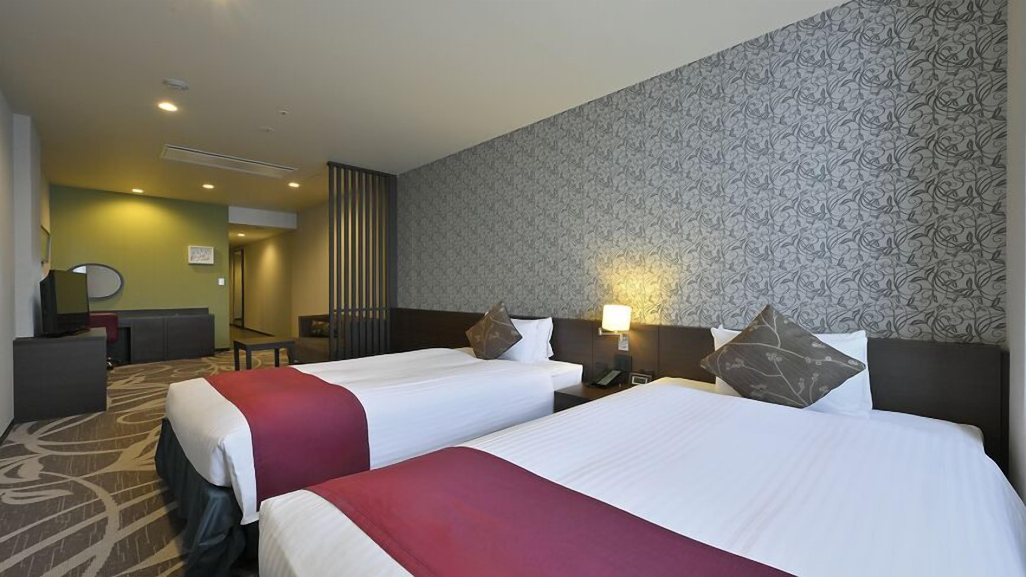 A hotel room with two beds, a desk, a chair, a TV, and a patterned wall.