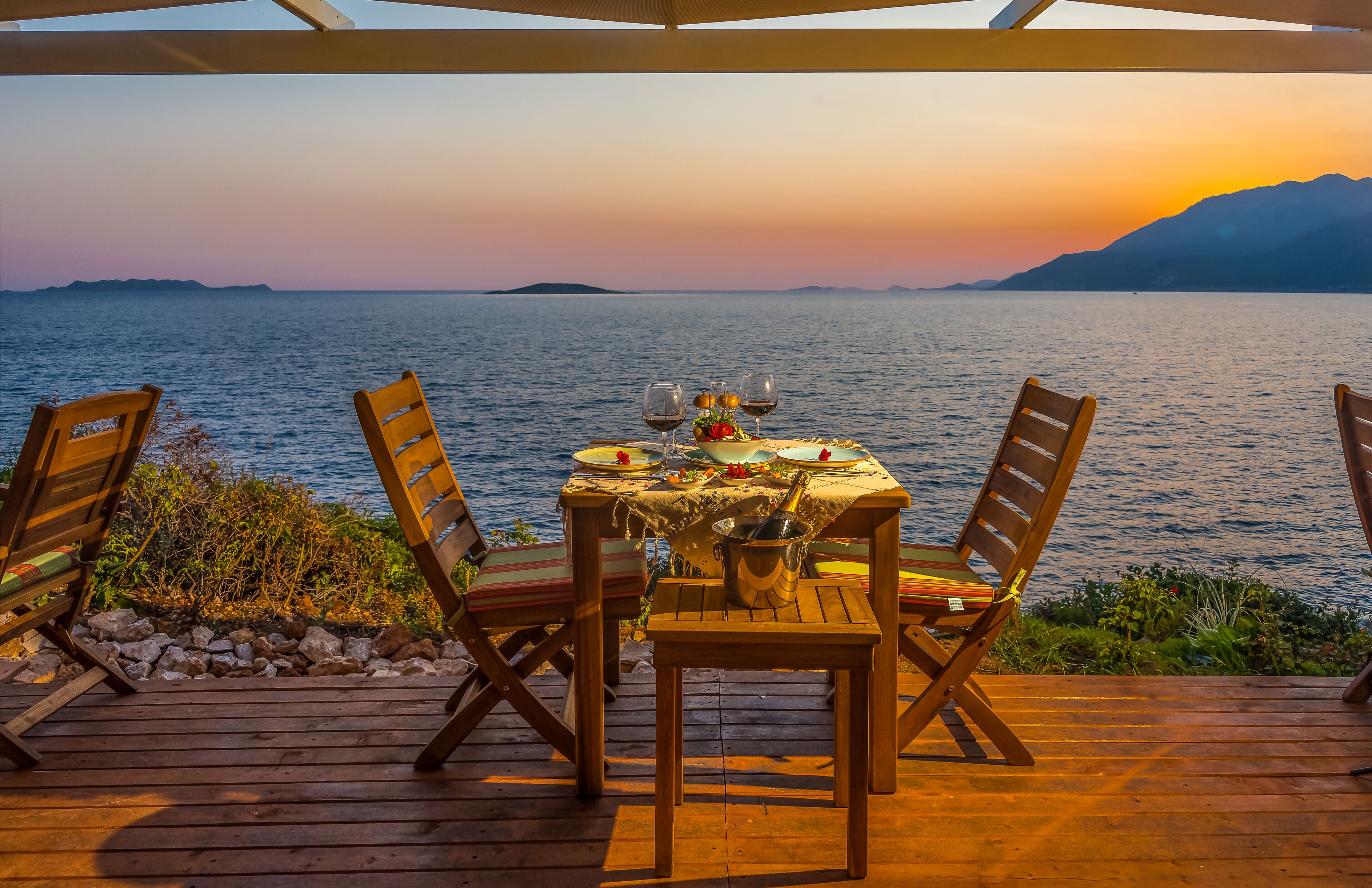 Una terraza de madera con una mesa dispuesta para dos, con vistas a un mar tranquilo y montañas distantes.