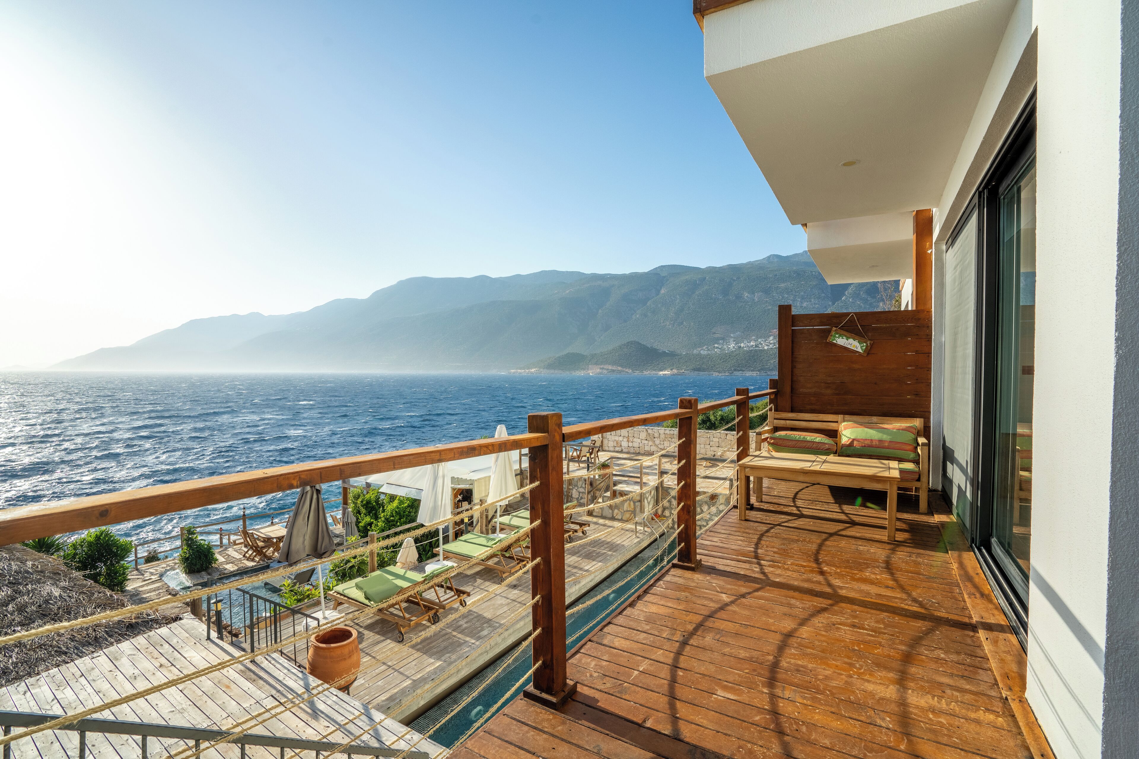 A balcony with a wooden deck, outdoor seating, and a view of the sea and mountains.