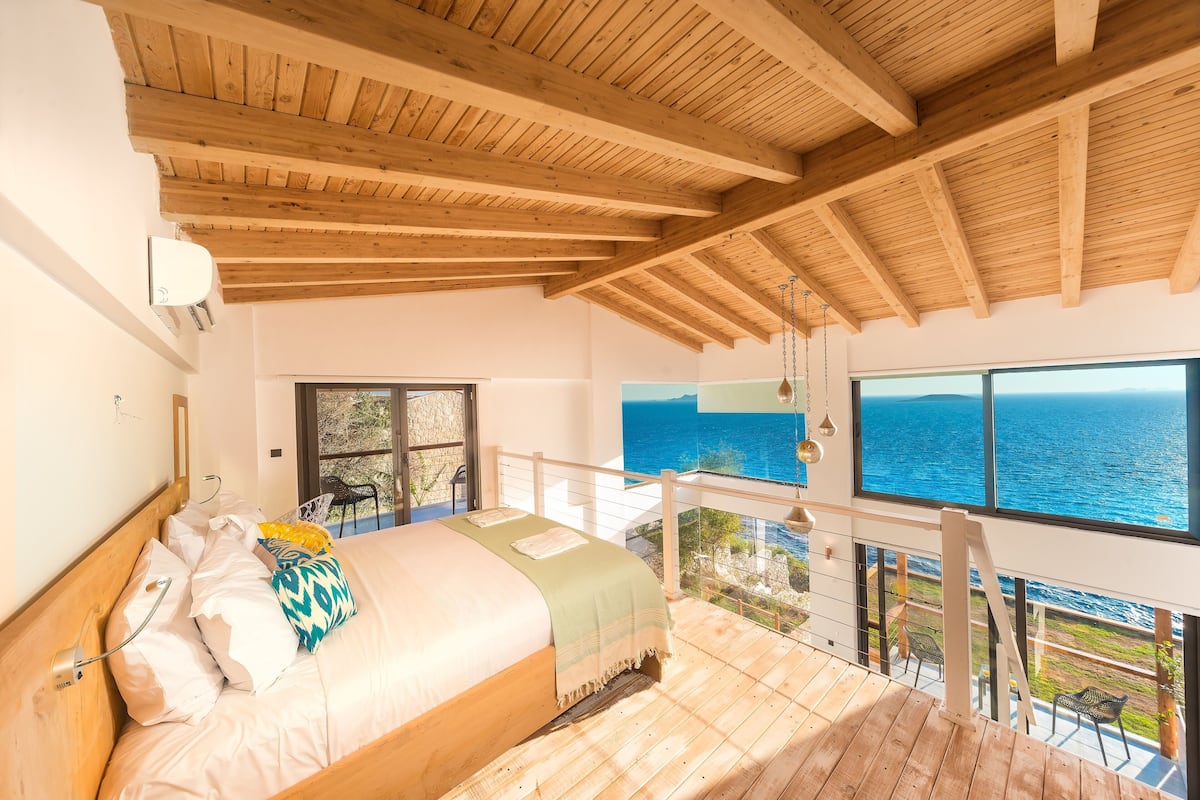 Un dormitorio con techo de madera, una cama, un ventanal con vista al mar y un balcón.
