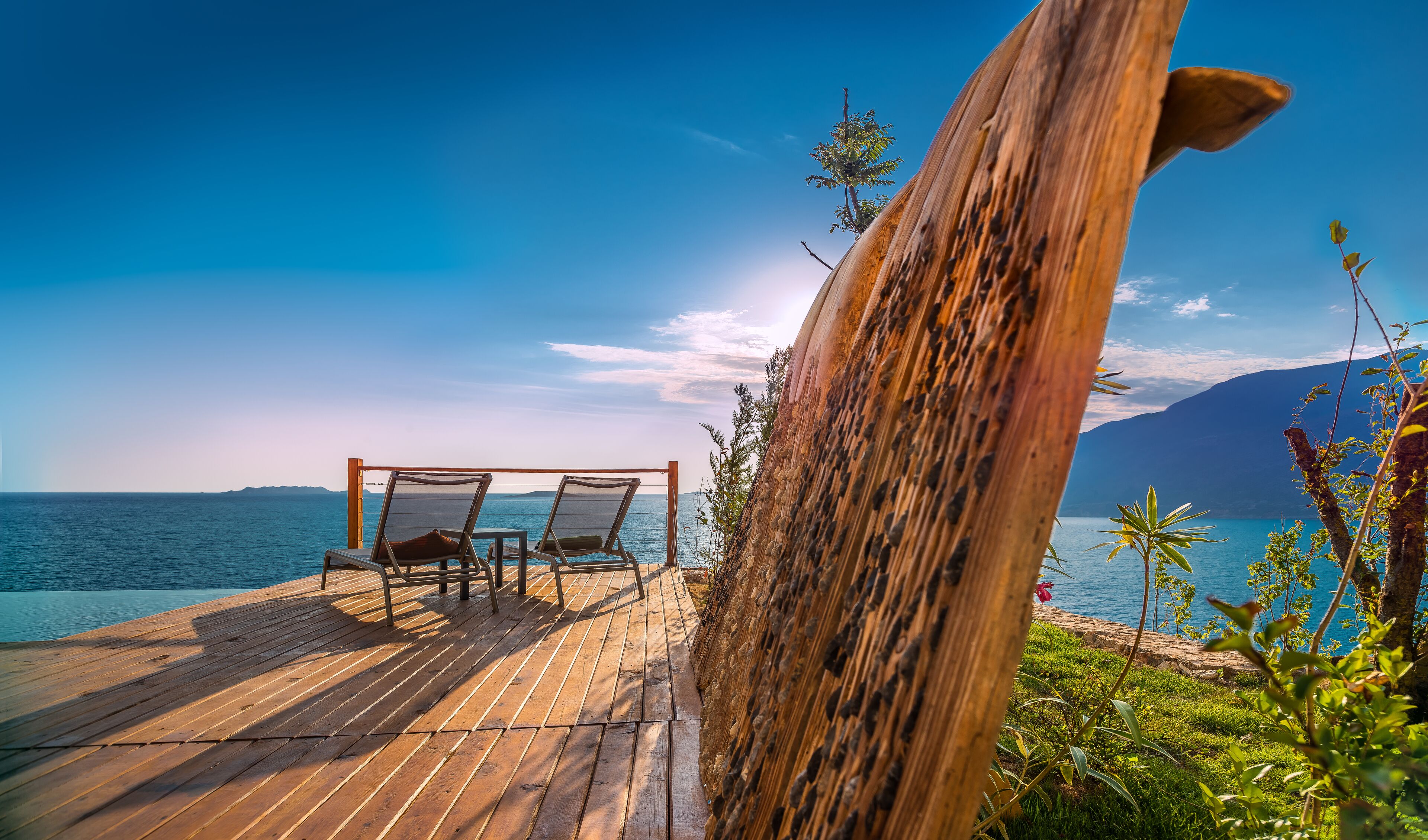 A wooden deck with two chairs and a table overlooking a calm sea and distant mountains.