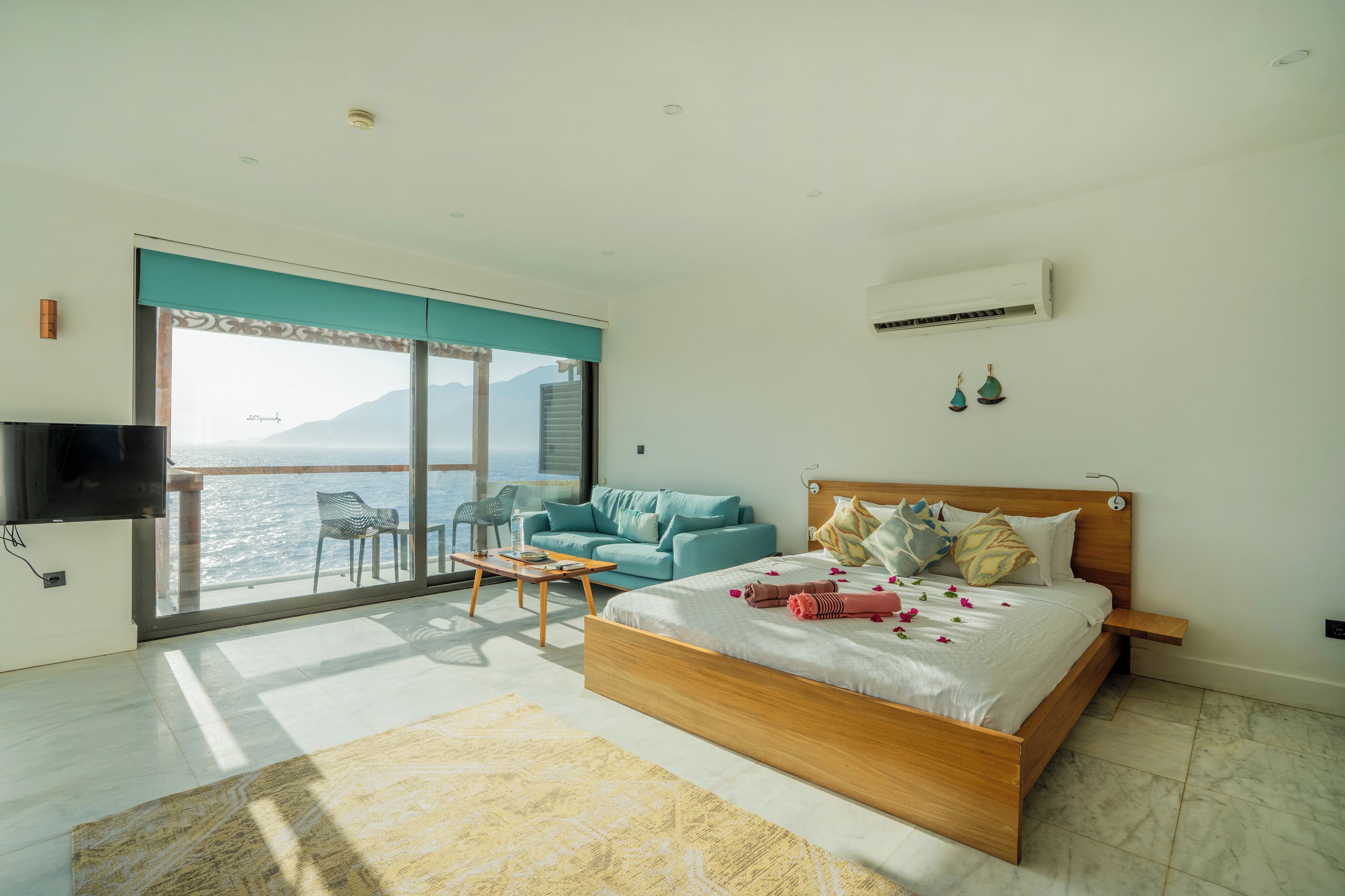 A modern bedroom with a large bed, a television, a sofa, and a balcony with a view of the sea.