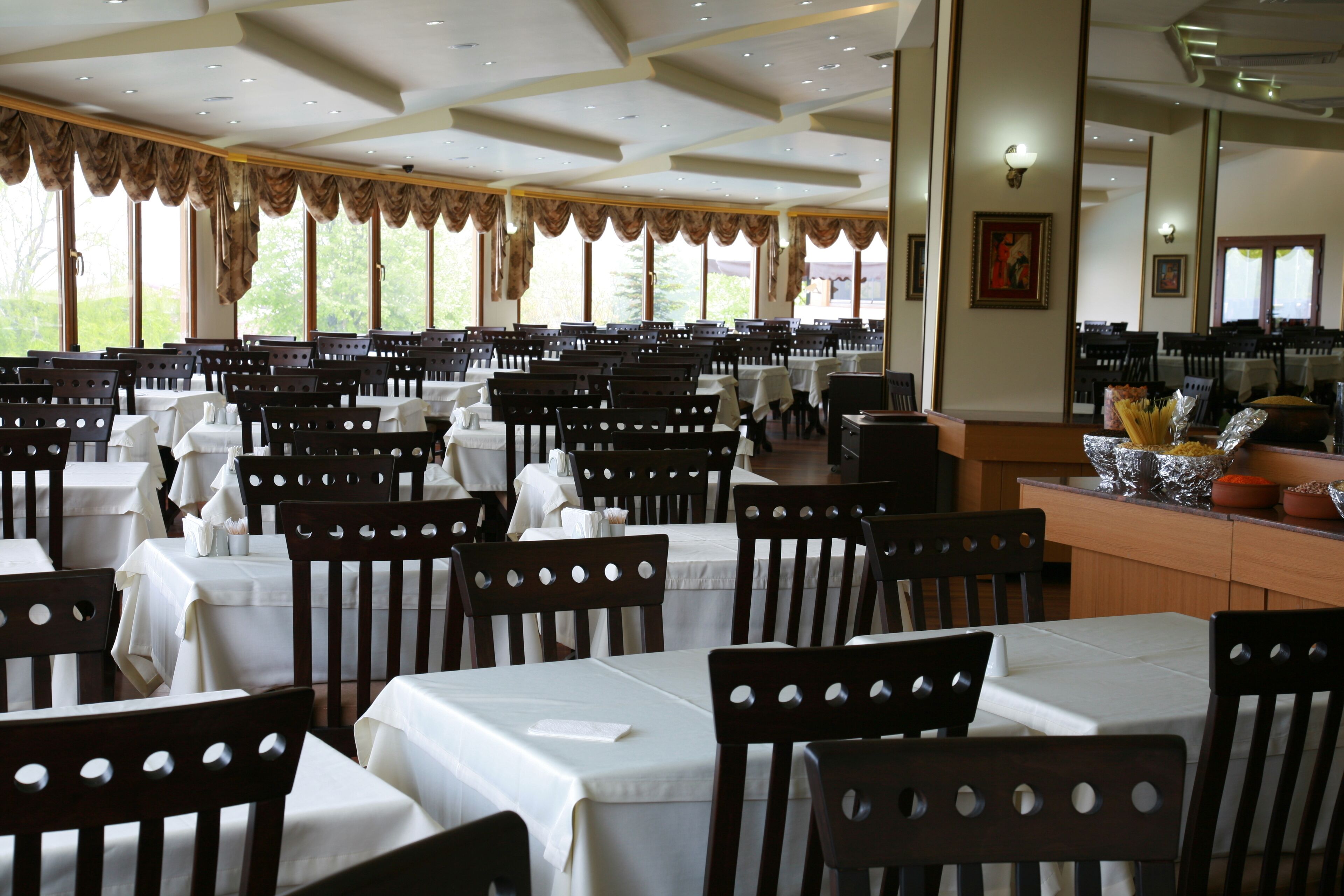Un restaurante con varias mesas y sillas, ventanas amplias y una barra con alimentos.