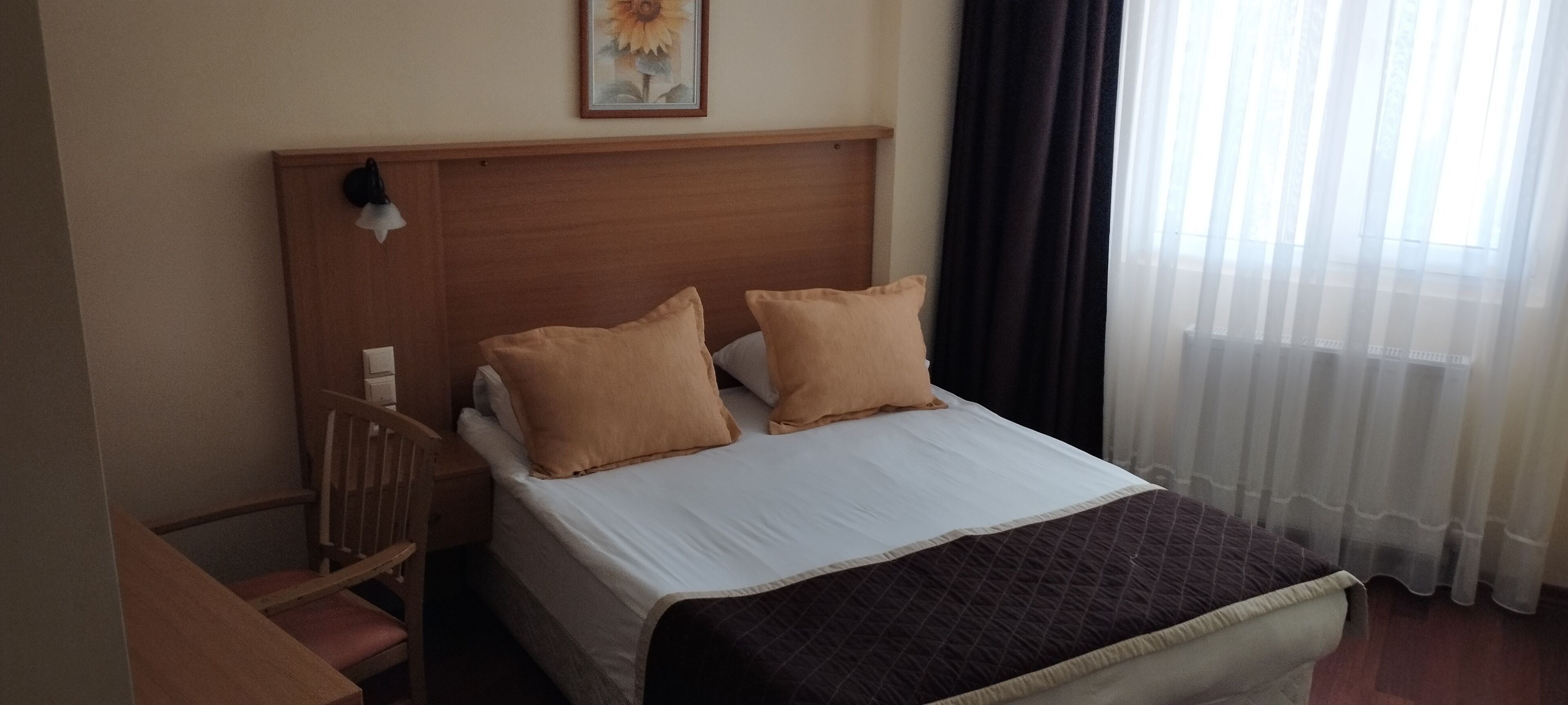 Une chambre d’hôtel avec une tête de lit en bois, deux oreillers beige, un couvre-lit blanc, une courtepointe marron foncé, une table de chevet en bois, une lampe et un tableau encadré au mur.