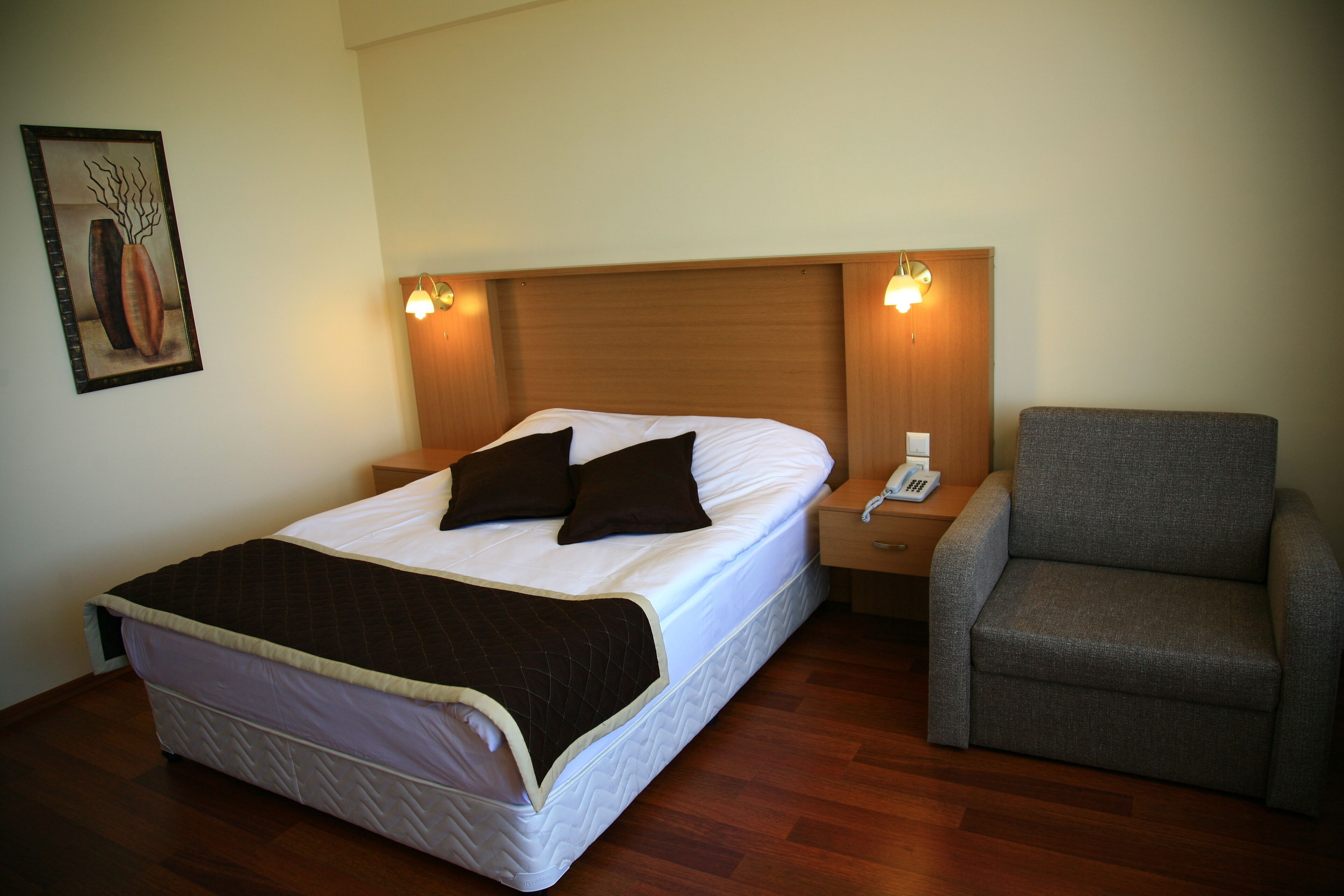 A neatly made bed with a brown blanket, a grey armchair, and a bedside table with a phone.