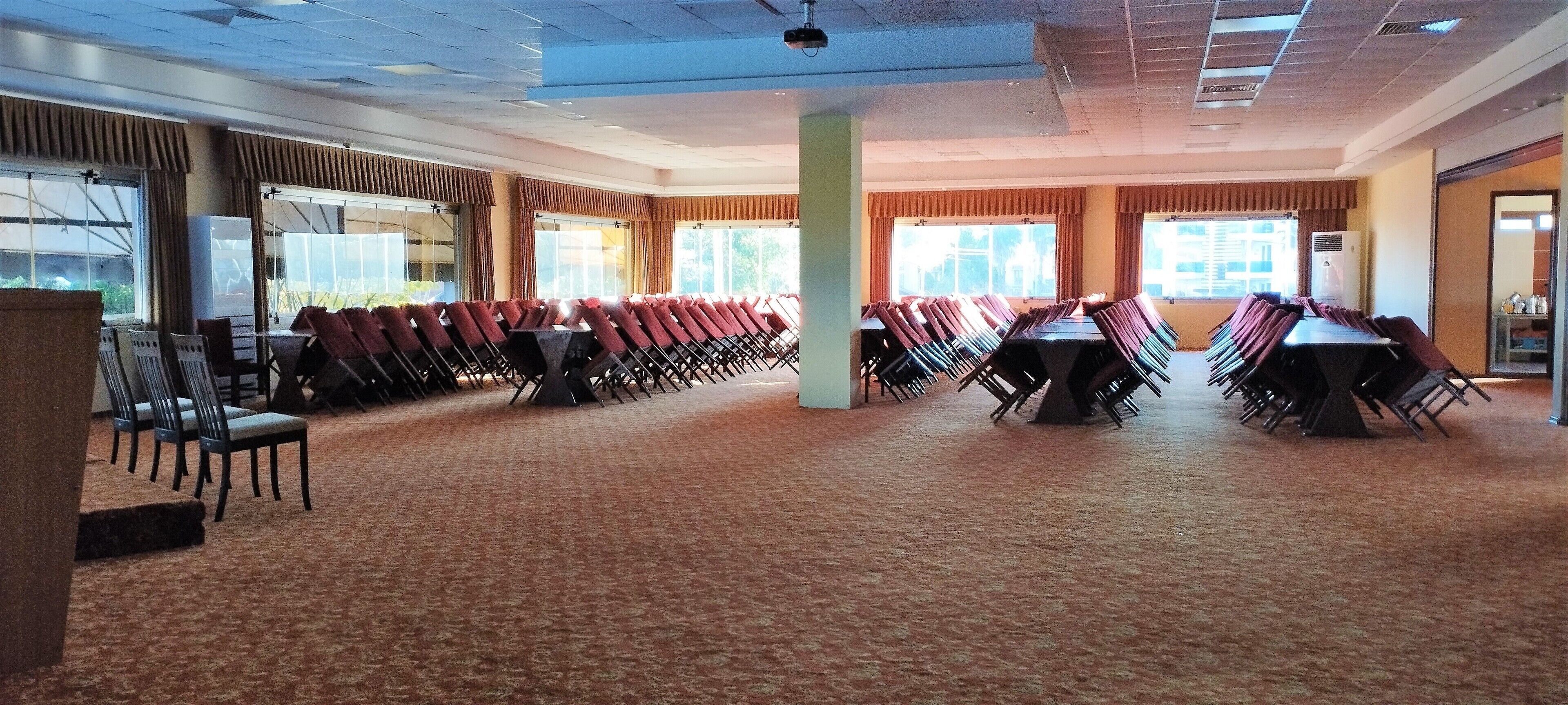 Un amplio salón de conferencias con filas de sillas y mesas, ventanas grandes y piso alfombrado.