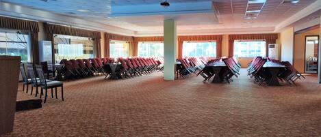 Un amplio salón de conferencias con filas de sillas y mesas, ventanas grandes y piso alfombrado.