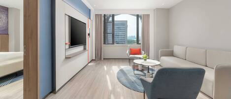 A modern hotel room with a large flat-screen TV, a sofa, a chair, and a small table with a vase of flowers.