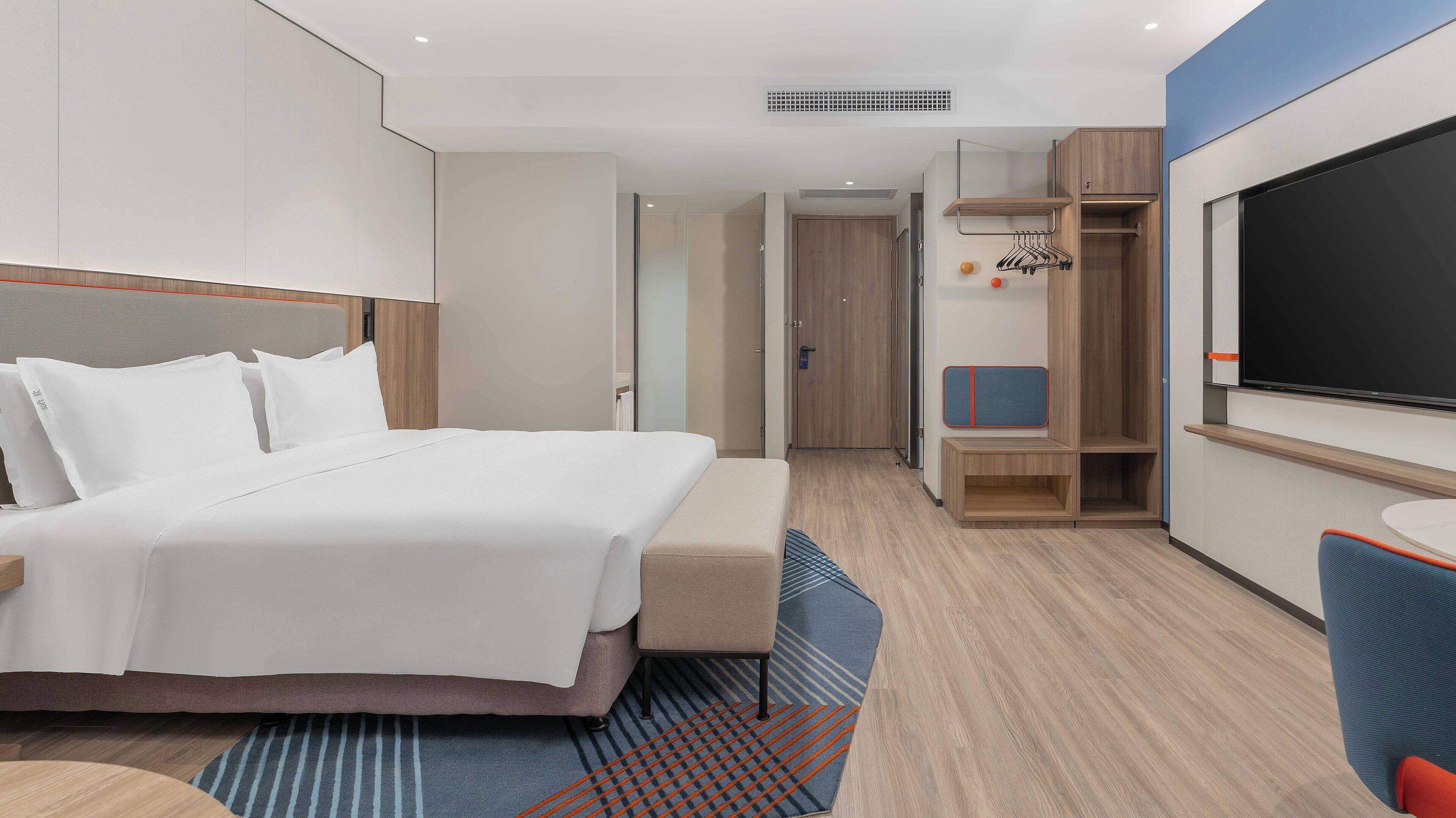 A modern hotel room with a large bed, a TV, a seating area, and a wardrobe.