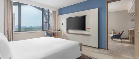 A modern hotel room with a large bed, a flat-screen TV mounted on the wall, a small table with a chair, and a view of the cityscape through large windows.