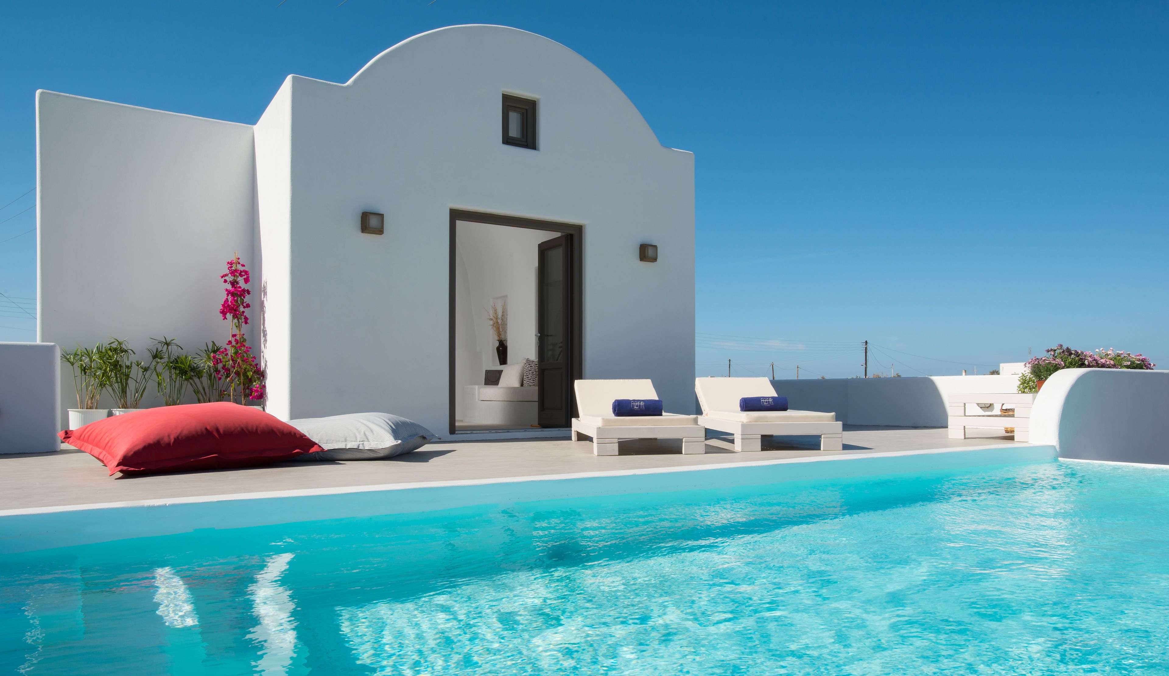 Un área moderna junto a la piscina con una pileta azul cristalina, sillas de descanso blancas y un edificio blanco con una puerta.