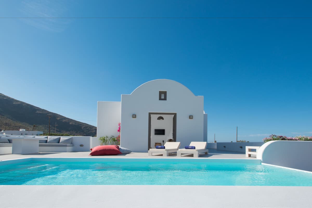 A modern pool area with white walls, a clear blue sky, and a mountain in the background.