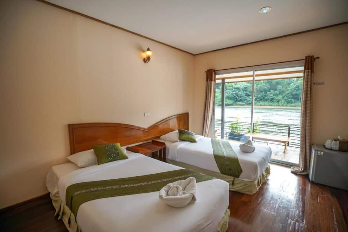 A hotel room with two beds, a nightstand, a lamp, a view of greenery outside, and a small table with items on it.