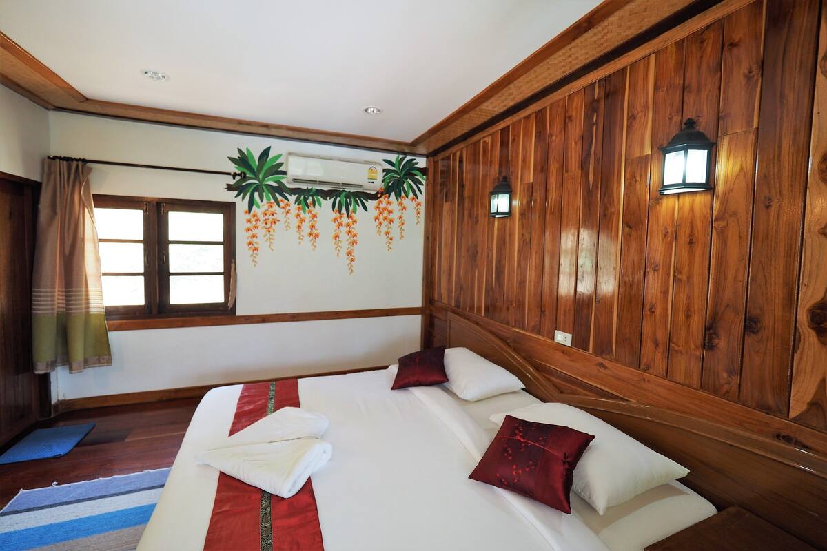 A hotel room with a wooden paneled wall, a large bed with white bedding and red pillows, a window with curtains, and a wall-mounted air conditioner.