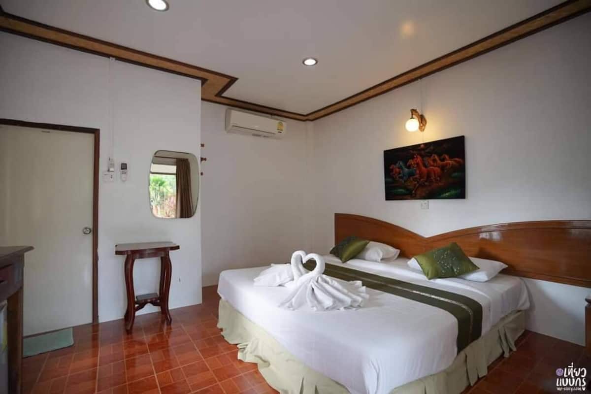 A hotel room with a bed, a bedside table, a mirror, an air conditioning unit, and a painting on the wall.