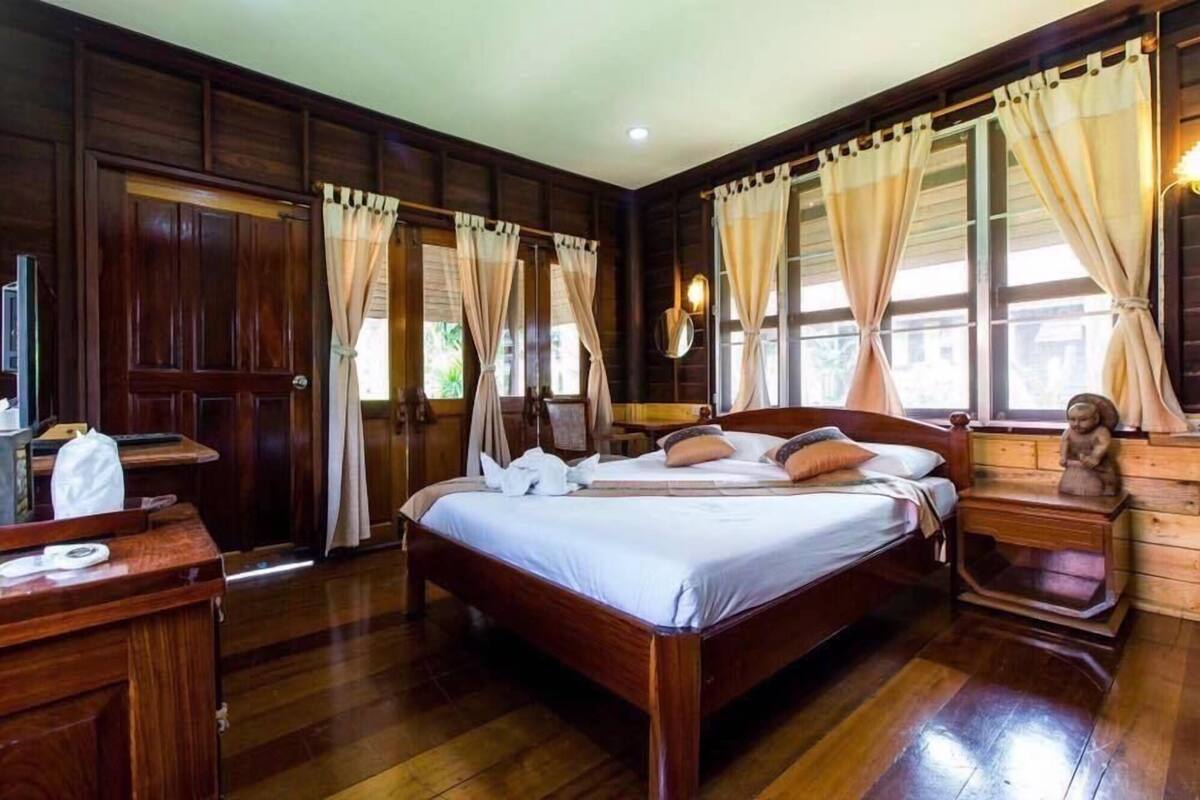 A wooden-floored bedroom with a large bed, curtains, a bedside table, and a nightstand.
