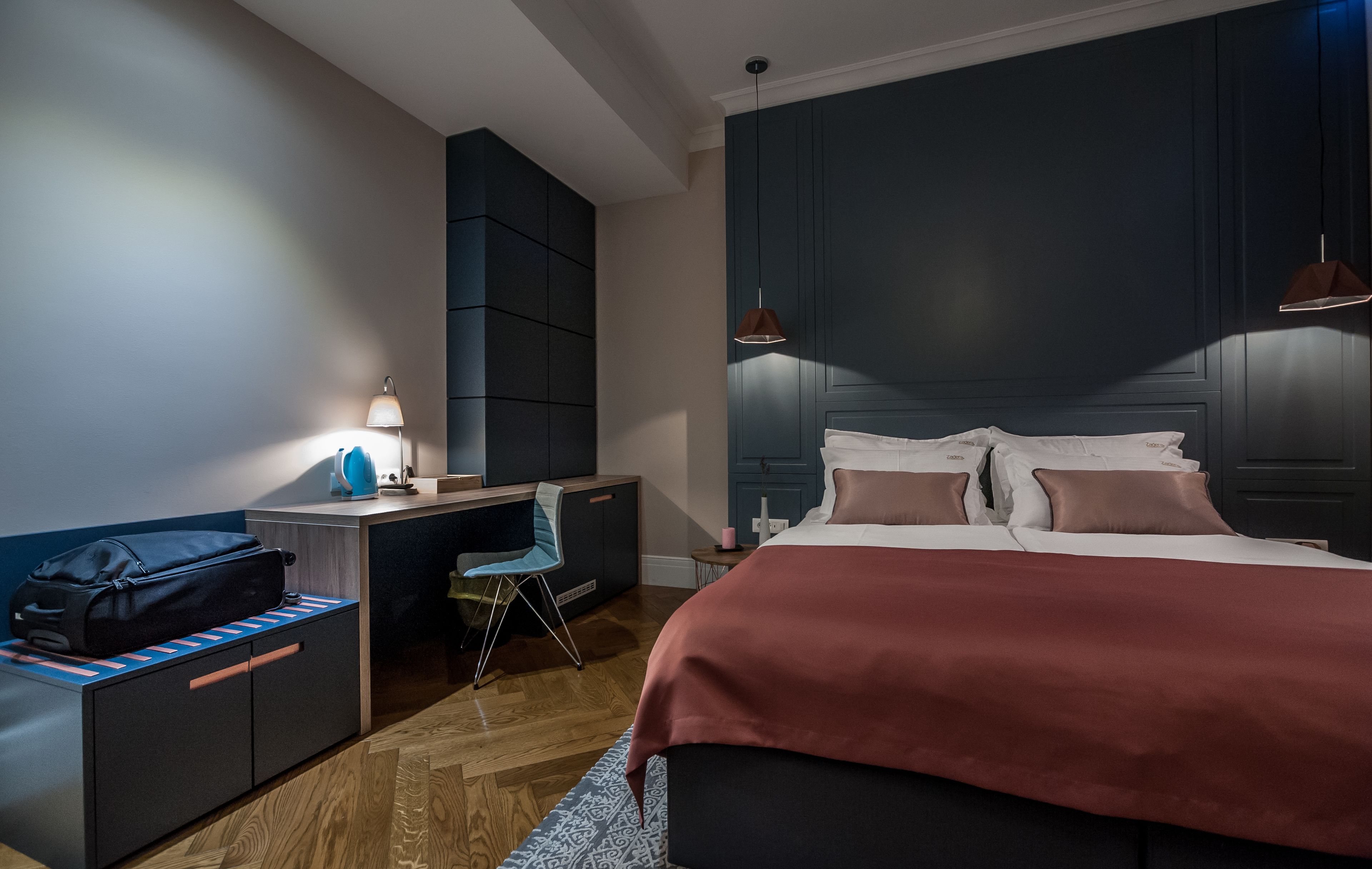 A modern hotel room with a large bed, a desk with a chair, a wardrobe, and a hanging light fixture.