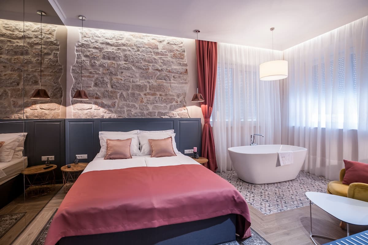 A modern hotel room with a large bed, a bathtub, a stone accent wall, and a view of the city.