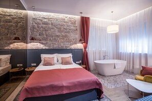 A modern hotel room with a large bed, a bathtub, a stone accent wall, and a view of the city. - Zadera Accommodation (Zadar)