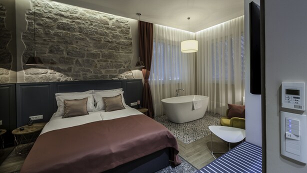 Une chambre moderne avec un mur d’accent en pierre, un grand lit, une baignoire et un coin salon séparé avec une petite table et une chaise.