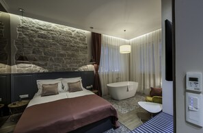 A modern bedroom with a stone accent wall, a large bed, a bathtub, and a separate sitting area with a small table and chair. - Zadera Accommodation (Zadar)