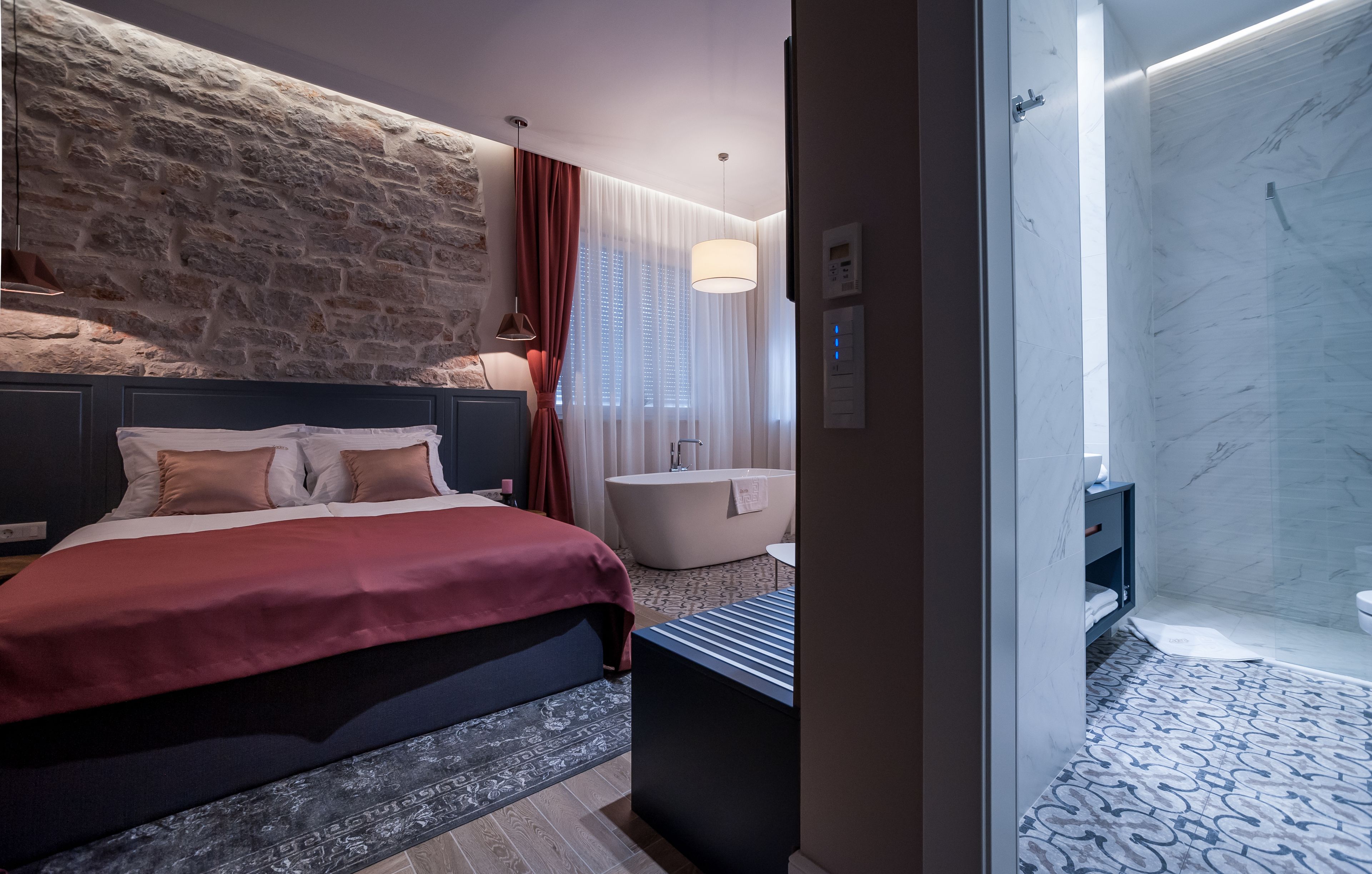 Una habitación de hotel moderna con pared de acento de piedra, una cama grande con ropa de cama roja, un baño separado con bañera, y piso con diseño.