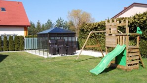 Un patio trasero con área de juegos, que incluye un tobogán y una estructura de escalada, un espacio de estar cubierto, y una casa con techo rojo.