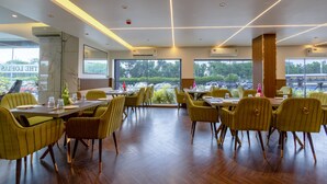 Un restaurante con sillas amarillas y una mesa de madera, amplias ventanas con vista a árboles y coches, y un letrero que dice "THE LOUNGE".