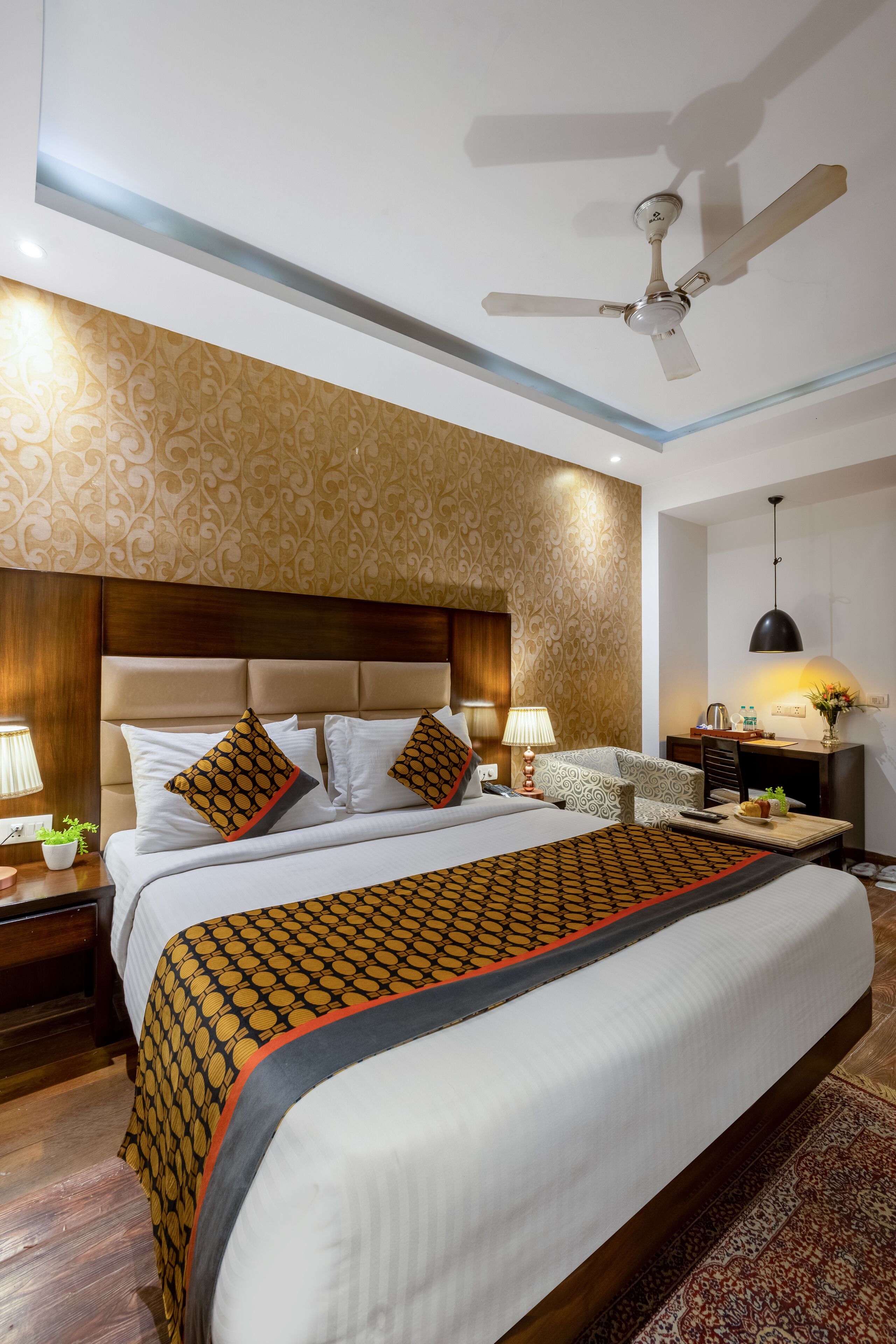 A hotel room with a large bed, a ceiling fan, a nightstand with a lamp, and a small table with a vase of flowers.